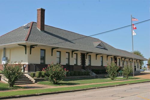 Historic building with a sloped roof, surrounded by greenery, featuring American and state flags on display.