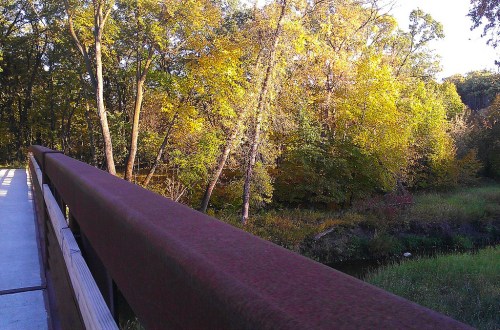 A wooden bridge with a rust-colored railing overlooks a vibrant autumn landscape with colorful trees and a stream.