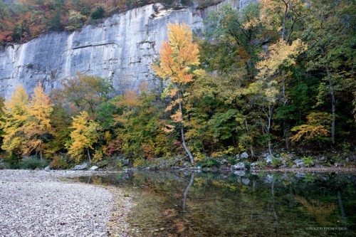 A serene riverbank with colorful autumn trees reflecting on the water, against a rocky cliff backdrop.