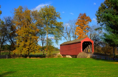 A red covered bridge surrounded by colorful autumn trees and a green field under a clear blue sky.
