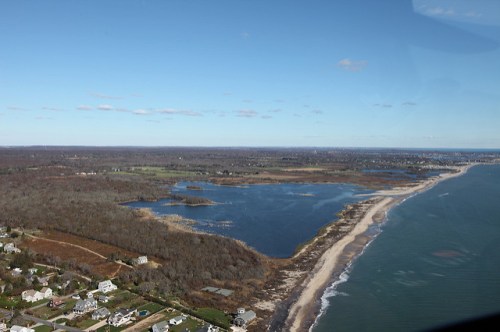 Aerial view of a coastal landscape featuring a beach, water bodies, and wooded areas under a clear blue sky.