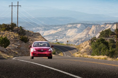 A red car drives along a winding road with mountains and power lines in the background under a clear sky.