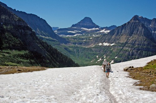 Two hikers walk on a snowy trail surrounded by mountains and lush green valleys under a clear blue sky.