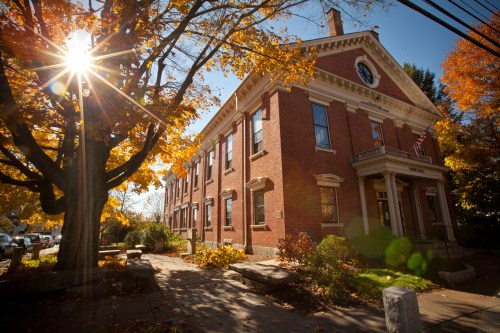 A brick building surrounded by autumn foliage, with sunlight shining through trees and a clear blue sky.