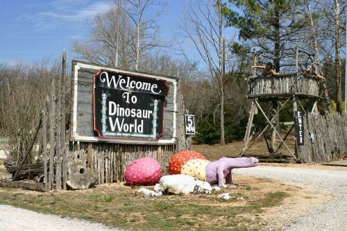 A rustic sign reading "Welcome to Dinosaur World" with colorful dinosaur sculptures and a lookout tower in the background.