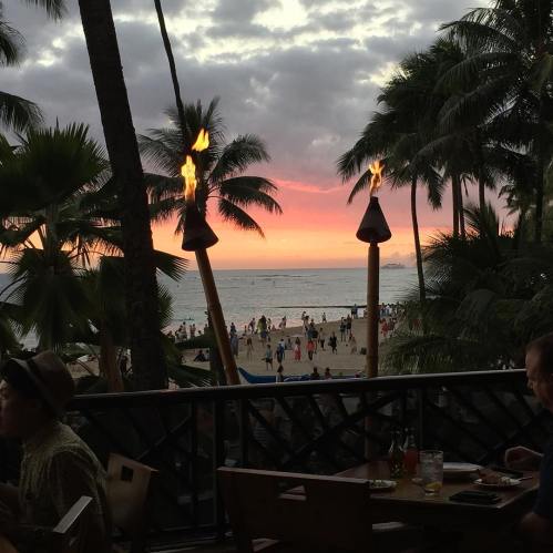 A tropical beach at sunset, with palm trees and tiki torches, and people enjoying the view in the foreground.
