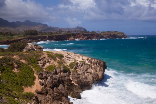A scenic coastal view with rocky cliffs, lush greenery, and waves crashing against the shore under a partly cloudy sky.