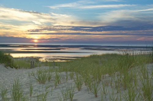 A serene beach scene at sunset, with sand dunes, grass, and a calm sea reflecting colorful clouds.