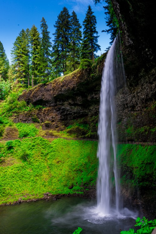 A tall waterfall cascades down rocky cliffs, surrounded by lush green trees and vibrant foliage under a clear blue sky.