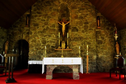 Interior of a stone chapel featuring an altar with candles, a crucifix, and statues, all set on a red carpet.