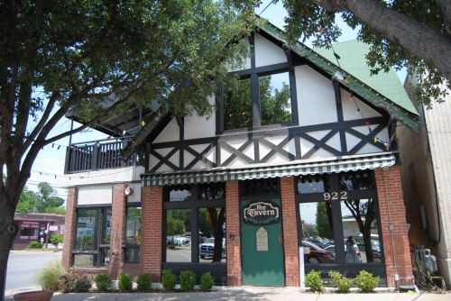 A charming two-story tavern with a green roof, brick facade, and decorative outdoor lights, surrounded by trees.
