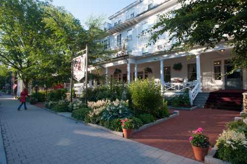 A charming hotel with a porch, surrounded by lush greenery and colorful flower pots, on a sunny day.