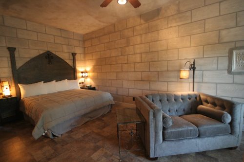 Cozy bedroom with a large bed, tufted gray sofa, and stone walls, featuring warm lighting and a rustic decor.