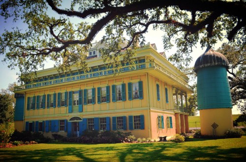 A colorful historic building with blue shutters and a green water tower, surrounded by lush greenery and flowers.