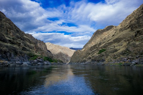 A serene river flows between steep, rocky mountains under a blue sky with scattered clouds.