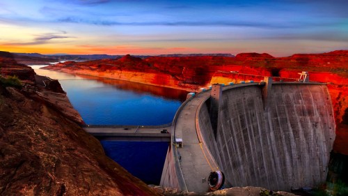 A panoramic view of a dam surrounded by red rock formations and a calm blue lake at sunset.