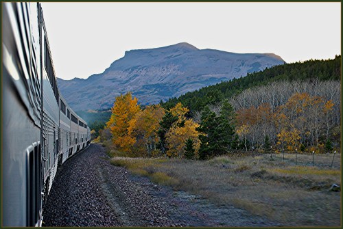 A train travels along a scenic route with autumn trees and a mountain in the background.