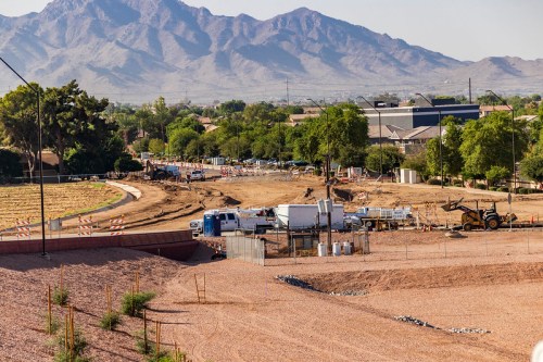Construction site with heavy machinery and vehicles, set against a backdrop of mountains and residential buildings.