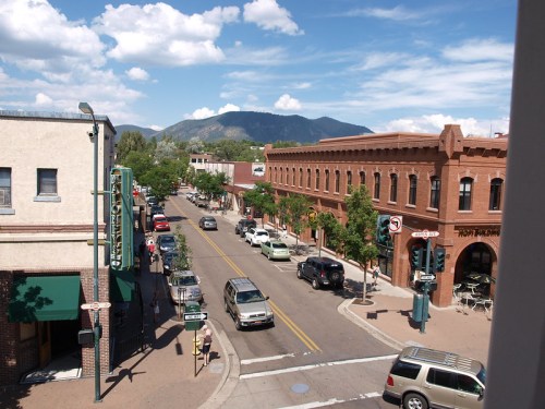 A bustling street scene with shops, cars, and mountains in the background under a blue sky with fluffy clouds.