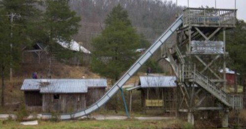 A tall water slide next to rustic buildings and trees, set in a rural landscape.
