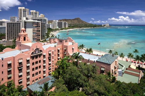 Aerial view of a pink hotel by the beach, with turquoise waters and city skyline in the background, under a clear blue sky.