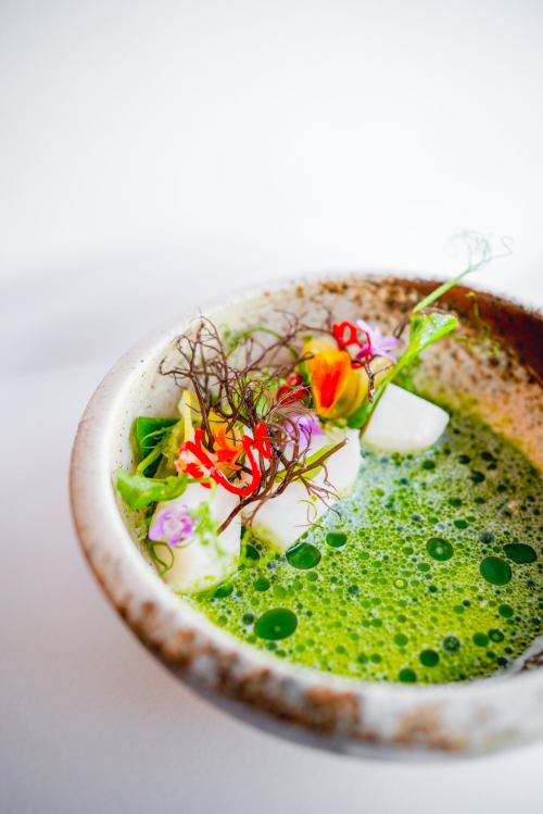 A bowl of vibrant green soup garnished with edible flowers, herbs, and small pieces of white ingredients.