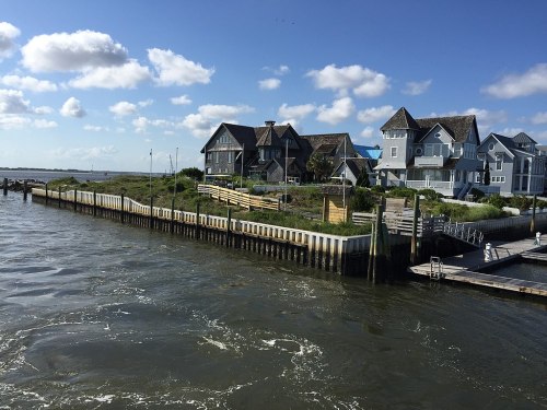 Coastal scene featuring houses on a grassy shore, with water and a cloudy blue sky in the background.