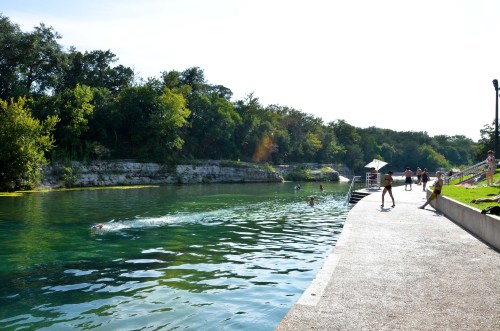 A serene river scene with people swimming and walking along a path, surrounded by lush greenery and bright sunlight.