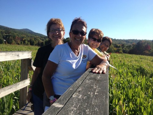 Four women smile and pose together on a wooden walkway, surrounded by green fields and a clear blue sky.