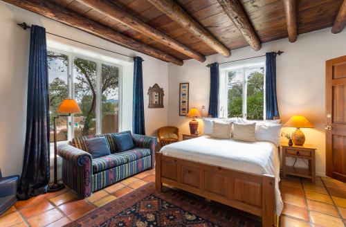 Cozy bedroom with a wooden bed, colorful couch, and large windows, featuring warm lighting and rustic decor.