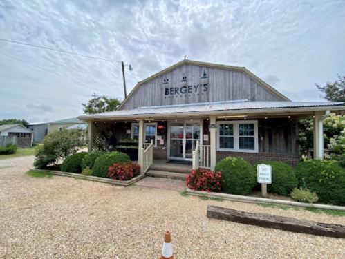 A rustic building with a sign reading "Bergey's" surrounded by flowers and gravel, under a cloudy sky.