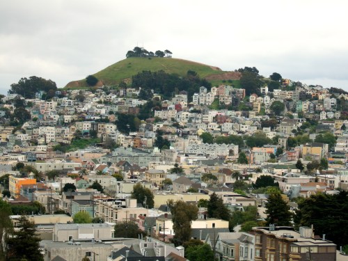 A view of a hilly neighborhood with colorful houses and greenery in the background under a cloudy sky.