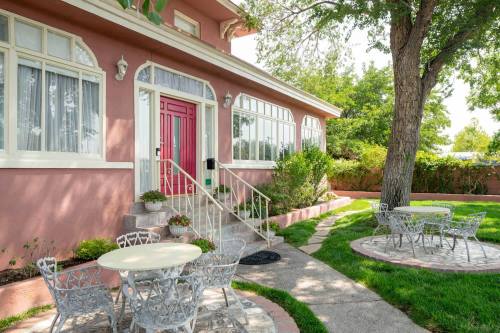 A charming pink house with a red door, surrounded by greenery and outdoor seating areas on a sunny day.