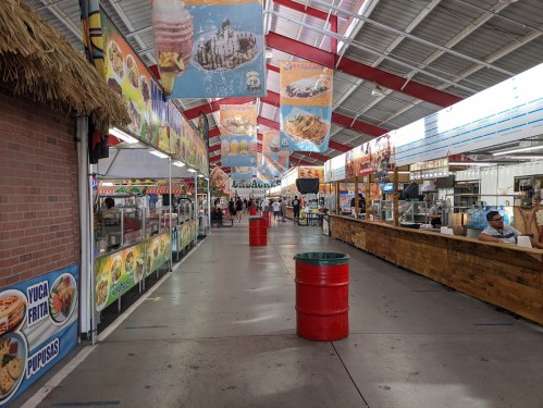 A vibrant food market with colorful banners, food stalls, and people walking down a spacious aisle.