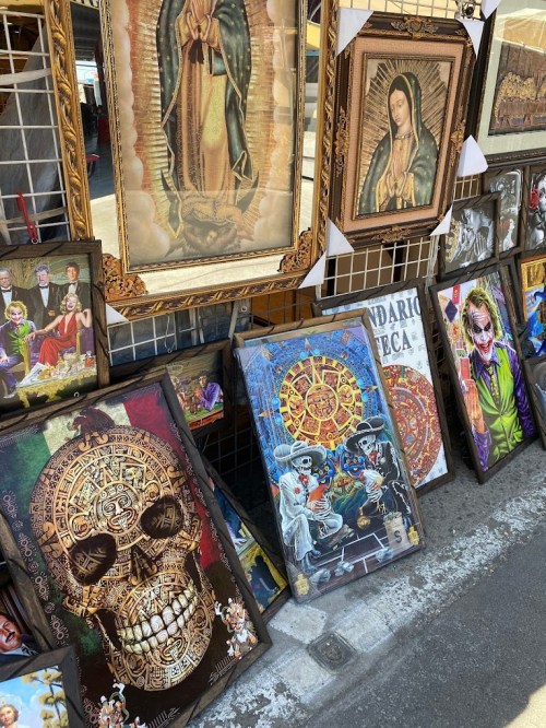A display of various framed artworks, including religious images, skull designs, and colorful characters, at a market.