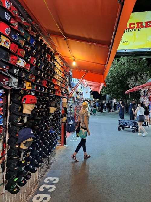 A person walks through a vibrant market aisle lined with colorful hats and stalls under orange canopies.