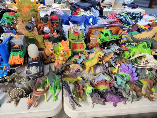A variety of colorful toy dinosaurs and vehicles displayed on a table, surrounded by clothing and other items.