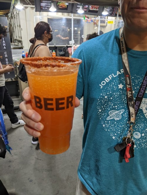 A person holds a large cup labeled "BEER" filled with a fizzy orange drink, rimmed with chili powder.