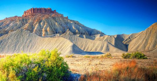 A rugged landscape featuring a prominent rocky hill, layered earth formations, and sparse vegetation under a clear blue sky.