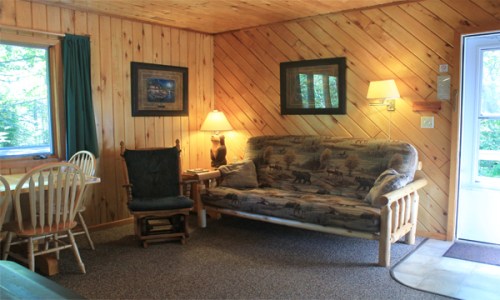 Cozy cabin interior with wooden walls, a sofa, chair, dining table, and warm lighting.