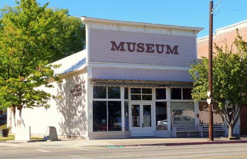 A small, light-colored museum building with a sign reading "MUSEUM" and trees nearby on a sunny day.