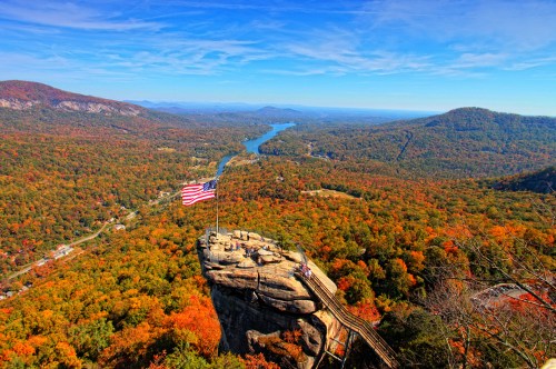 A scenic view of a valley with vibrant autumn foliage, a river, and an American flag atop a rocky outcrop.