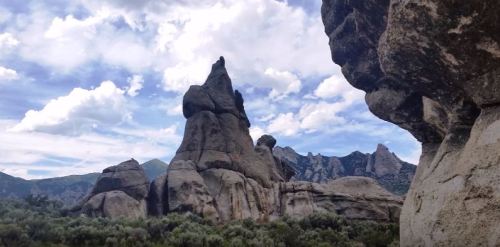A rocky landscape with towering formations under a cloudy sky, surrounded by greenery and distant mountains.