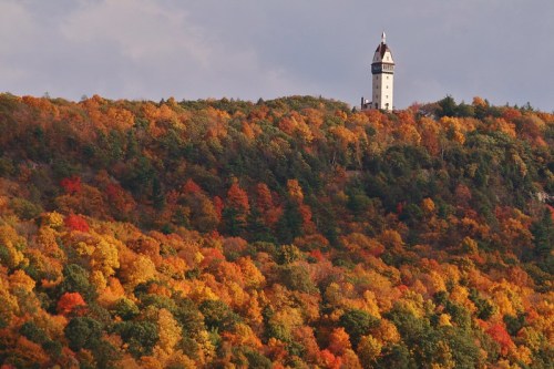 A tall tower stands atop a colorful hillside, surrounded by vibrant autumn foliage in shades of orange, red, and yellow.