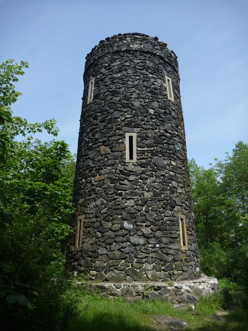 A tall, cylindrical stone tower surrounded by greenery under a clear blue sky.