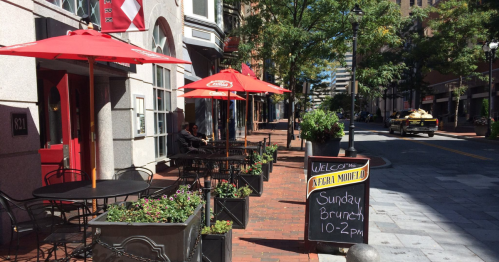 Outdoor café scene with red umbrellas, planters, and a chalkboard sign for Sunday brunch on a sunny street.