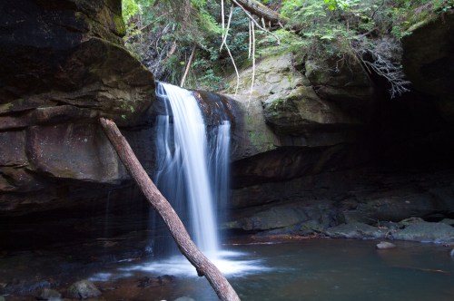 A serene waterfall cascades over rocks into a calm pool, surrounded by lush greenery and trees.