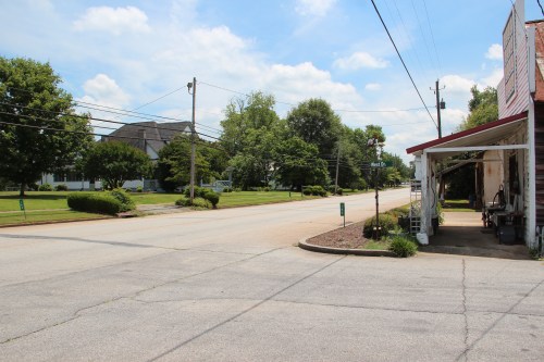 A quiet street scene with a view of a grassy area, trees, and a building on the right under a blue sky.