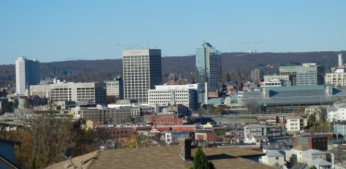 A panoramic view of a city skyline featuring tall buildings and a clear blue sky, with hills in the background.