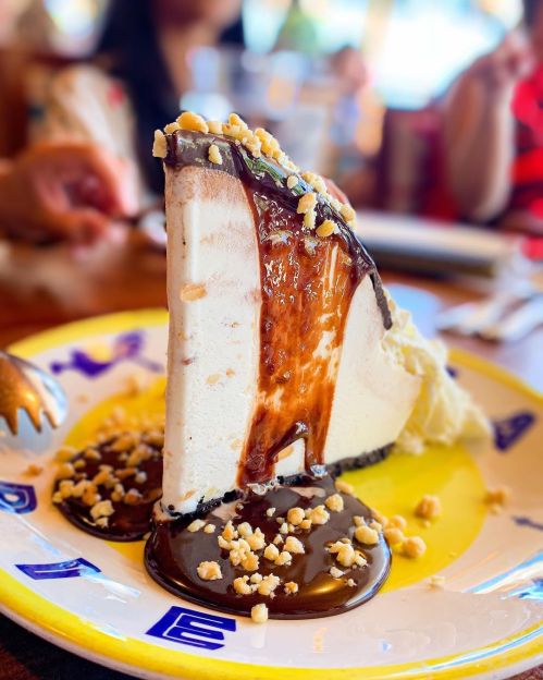 A slice of creamy dessert with chocolate sauce and crushed nuts on a colorful plate, with people in the background.